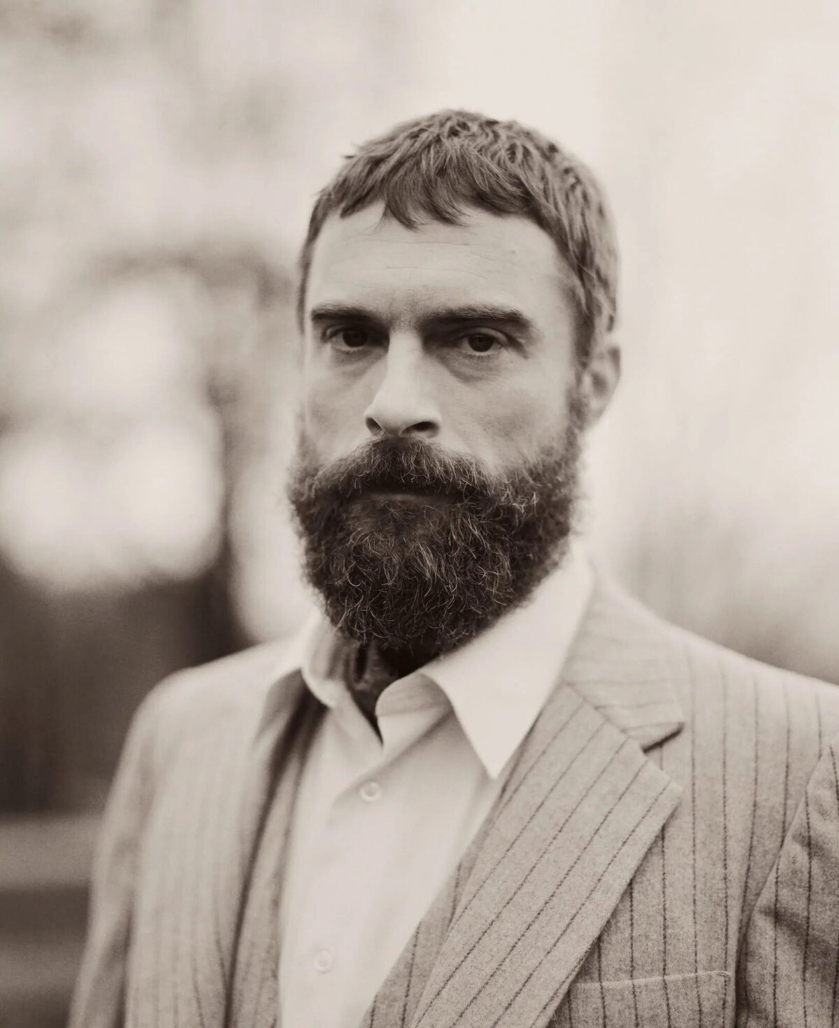 A portrait of a bearded man in a pinstripe suit, looking directly at the camera with an intense and distinguished gaze.