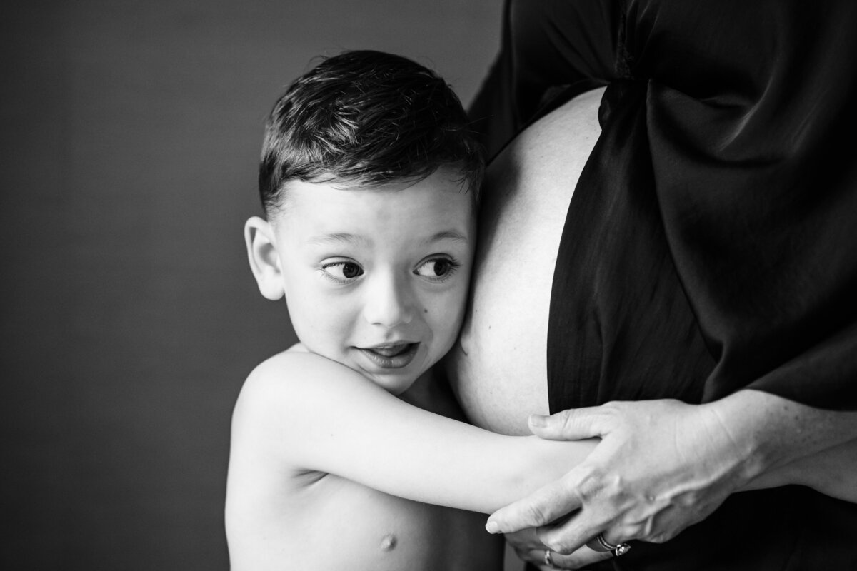 A close-up of a young boy hugging his mother’s pregnant belly. He looks up with a curious expression while she gently holds his hands.