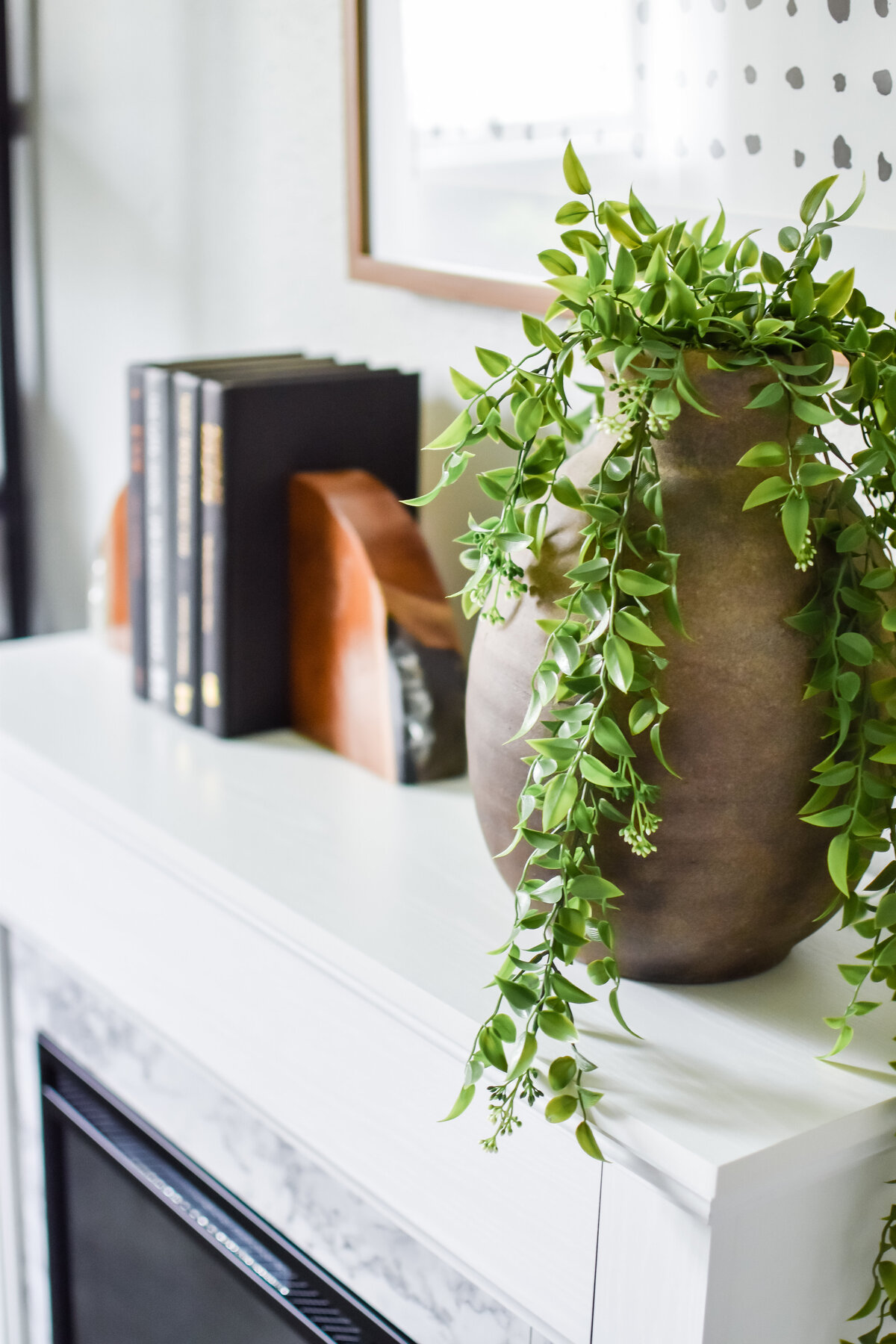 Plant stems hang out of a tall vase sitting on the corner of a fireplace mantle
