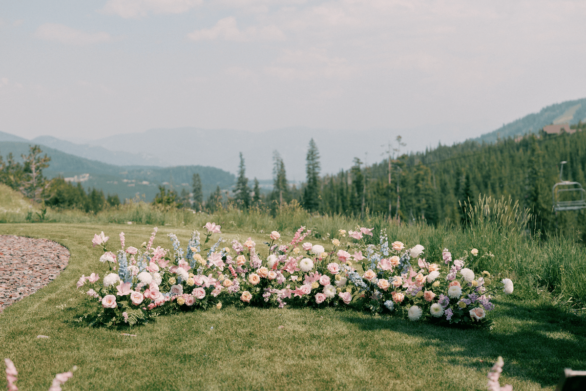 Ground floral arch with pastel roses, dahlias, and delphinium at a summer wedding at Moonlight Basin, framed by mountain views and natural greenery.
