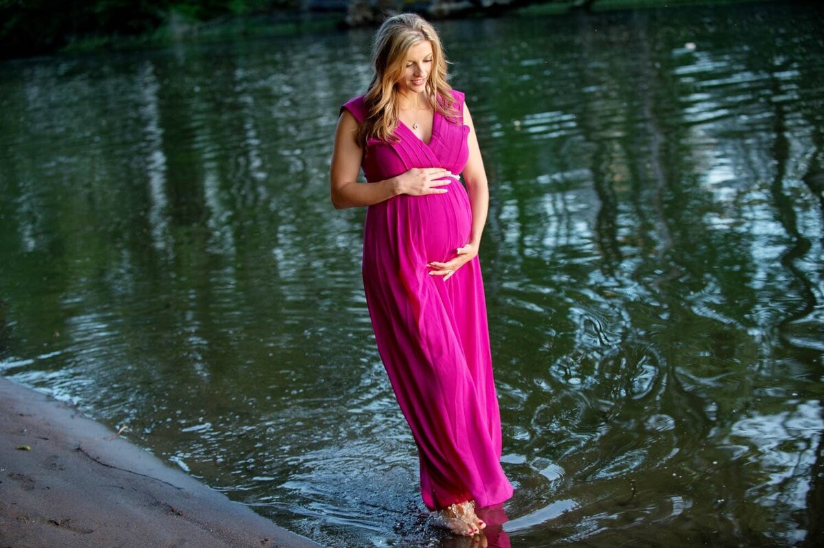 Beautiful expecting mother in a bright pink and long dress, dipping her feet in the lake water.
