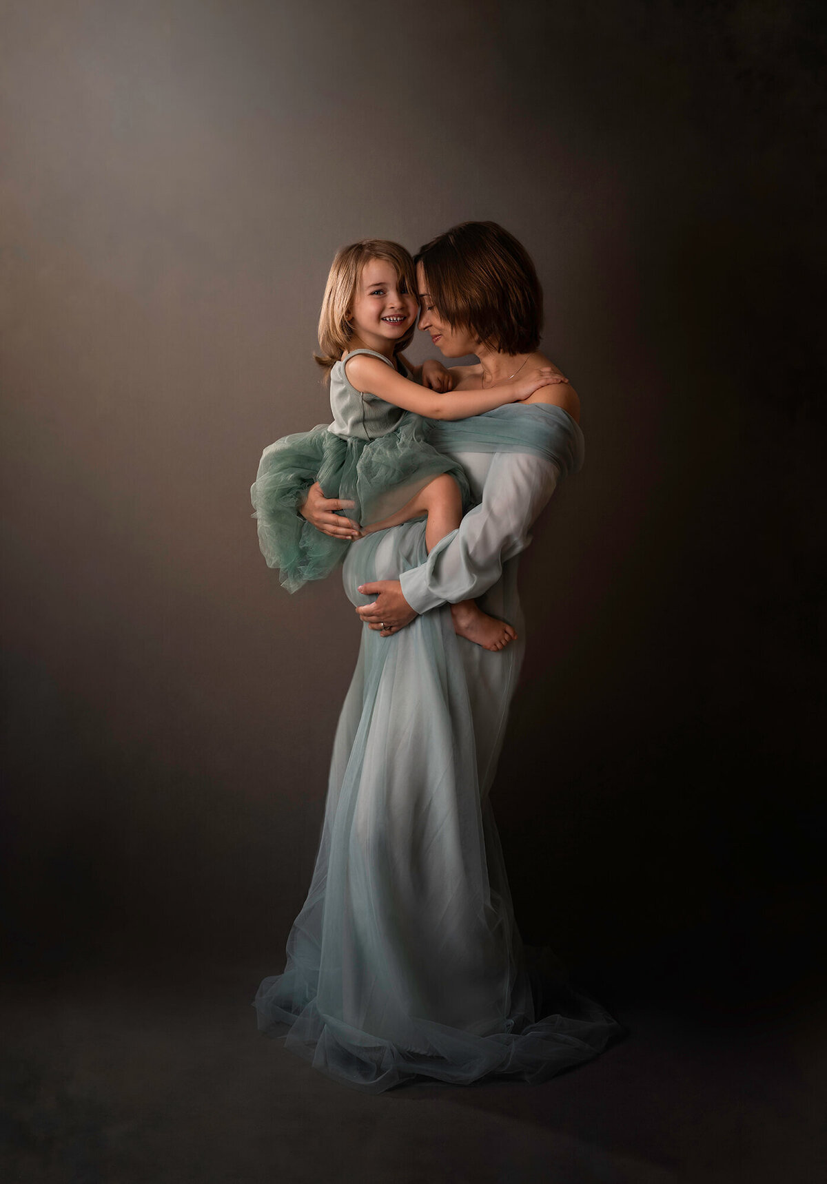 A beautiful pregnant mama in a long blue dress holds her daughter and cradles her baby bump