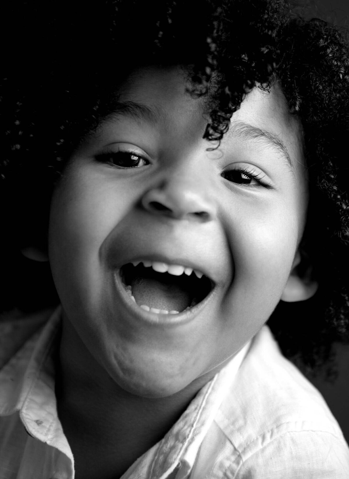 Black and white close-up of a child with curly hair, laughing joyfully. The expression conveys happiness and innocence, wearing a light-colored shirt.