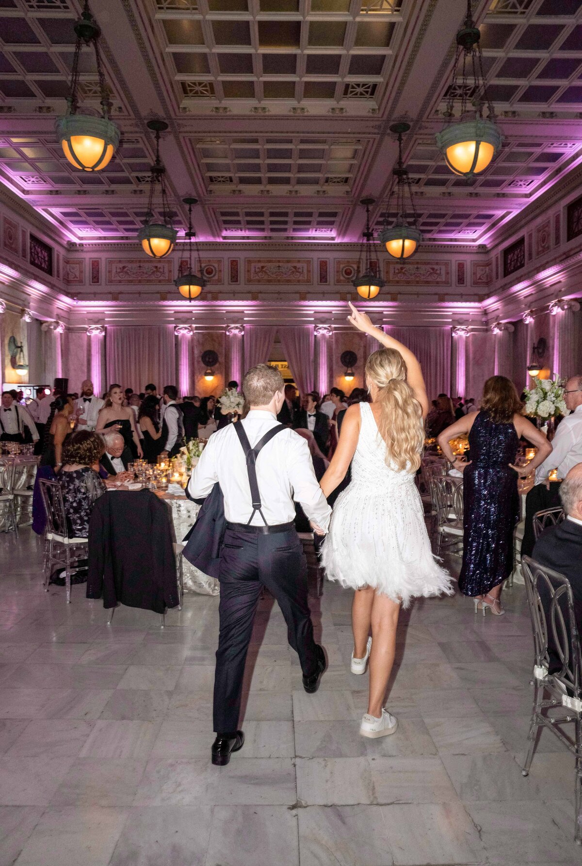 A couple in formal attire is joyfully dancing in a grand ballroom with elegant chandeliers and pink uplighting. The room is filled with guests seated at banquet tables. The atmosphere is festive and celebratory.