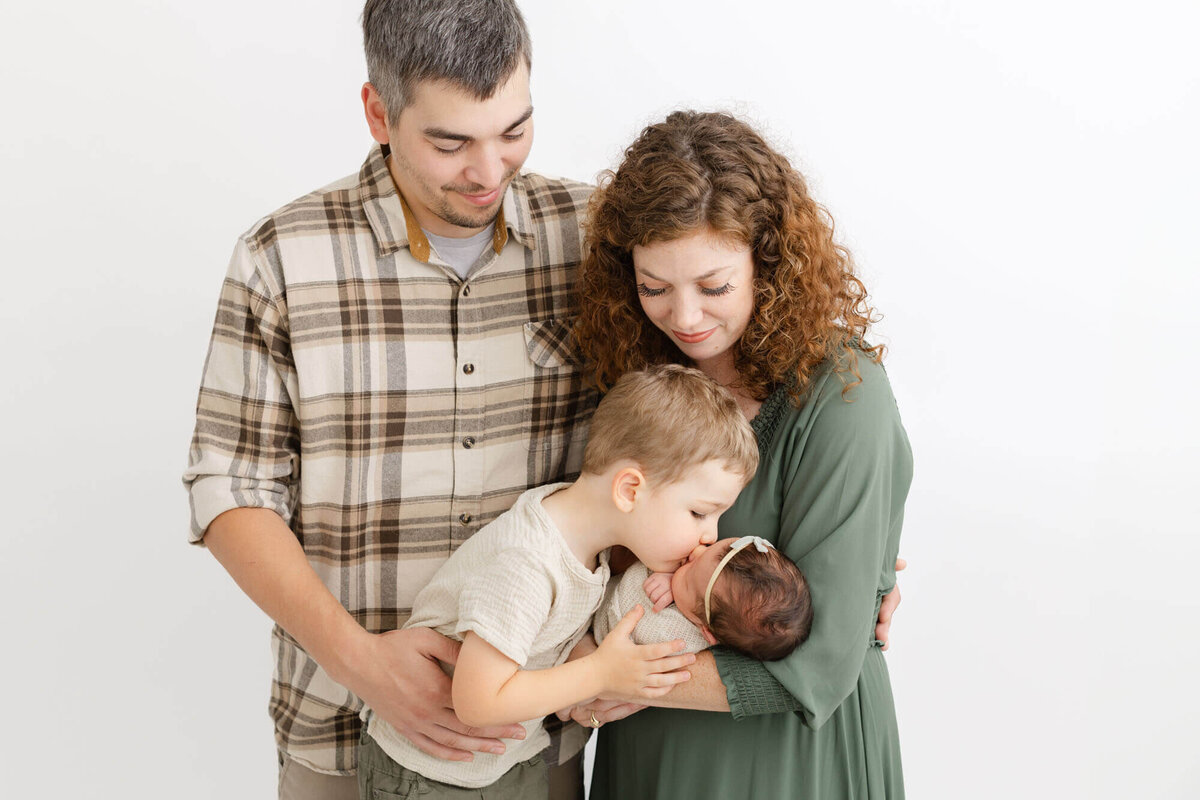 Mom holding baby in her arms while dad stands next to her and toddler brother is right in front of dad and leaning over baby and giving her a little kiss. Mom and Dad are looking down at the both of them and smiling.