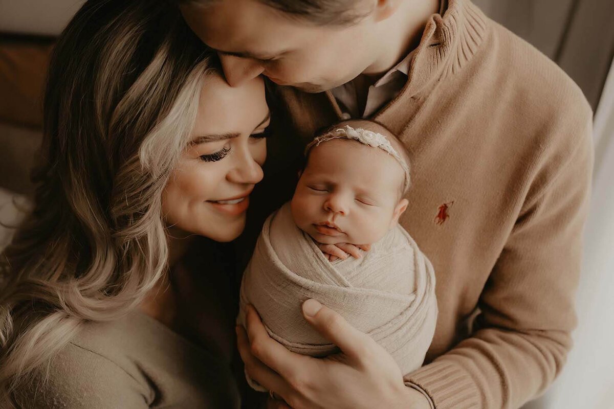 A dad kisses his wifes head while holding their newborn son while the mom looks at the baby