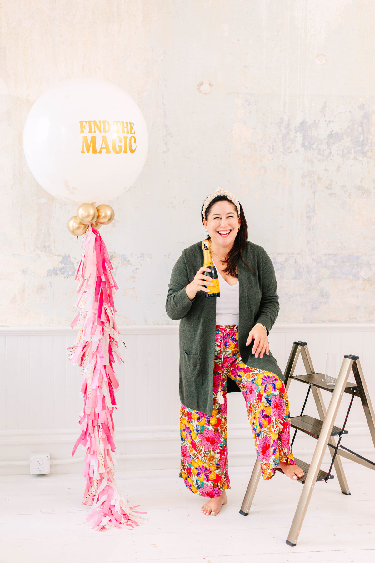 A woman in colorful floral pants and a dark green cardigan sits on a step ladder, laughing while holding a bottle. A large white balloon with "Find the Magic" written in gold and pink tassel decorations stands beside her.