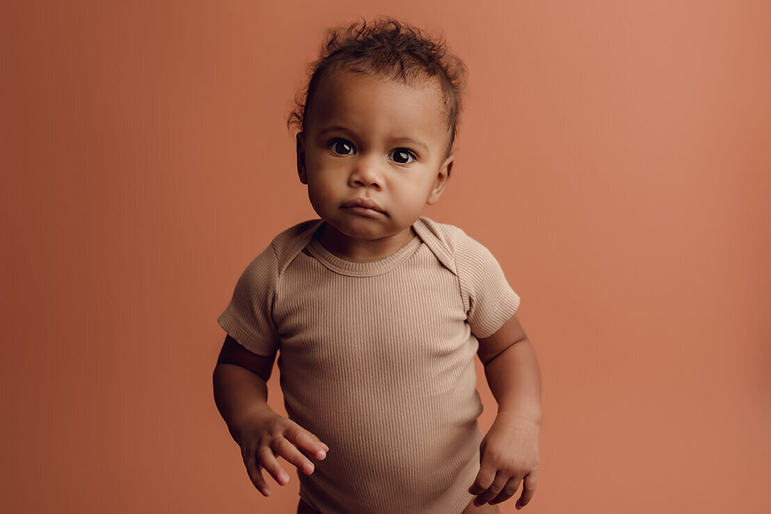 a one year old girl wearing a tan onesie on a tan backdrop looking at the camera