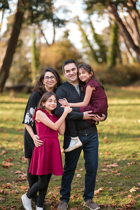 A family of four, dressed in fall colors, together in vibrant foliage of Lincoln Park in West Seattle. The sun filters through the trees at golden hour.  Casting a warm glow on the family. . This photo is a perfect example of fall family mini sessions with Heleyna Holmes Photography,