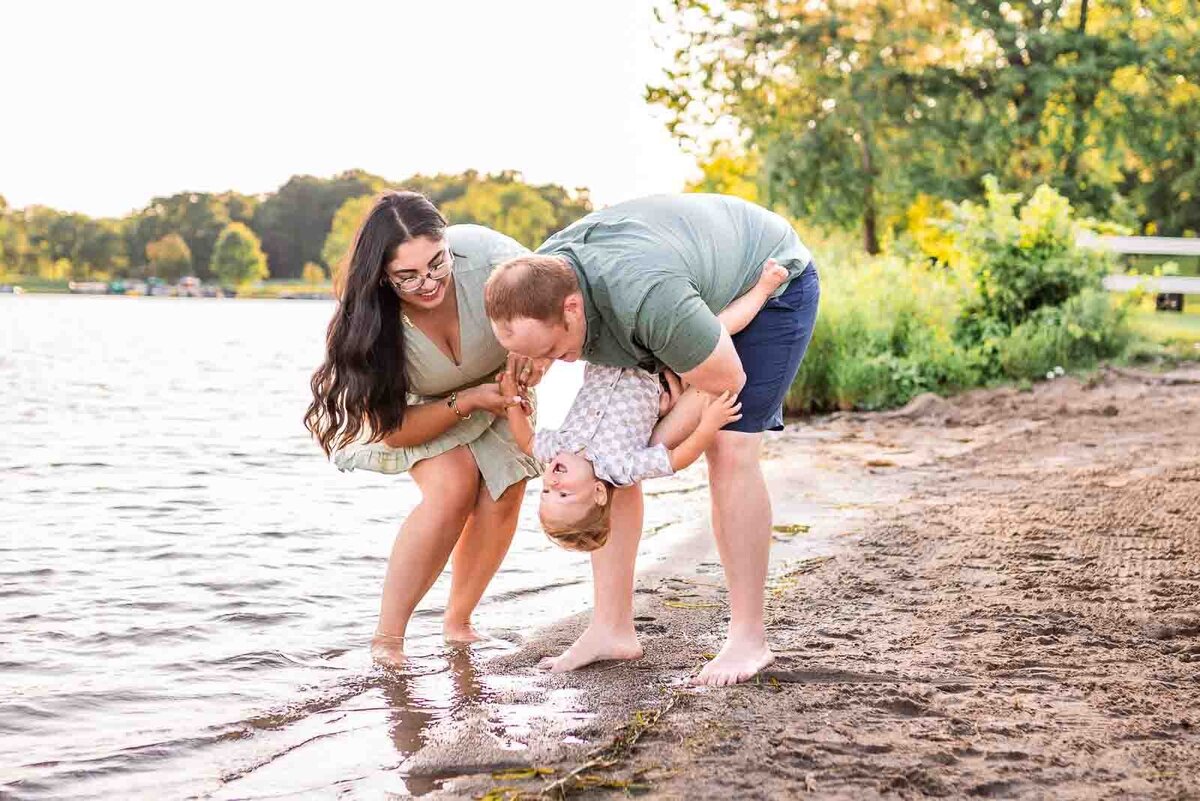 Dad holding toddler and tipping him backwards while mom is reaching towards baby smiling as they are dipping toes in the water at Kensington Metropark beach 