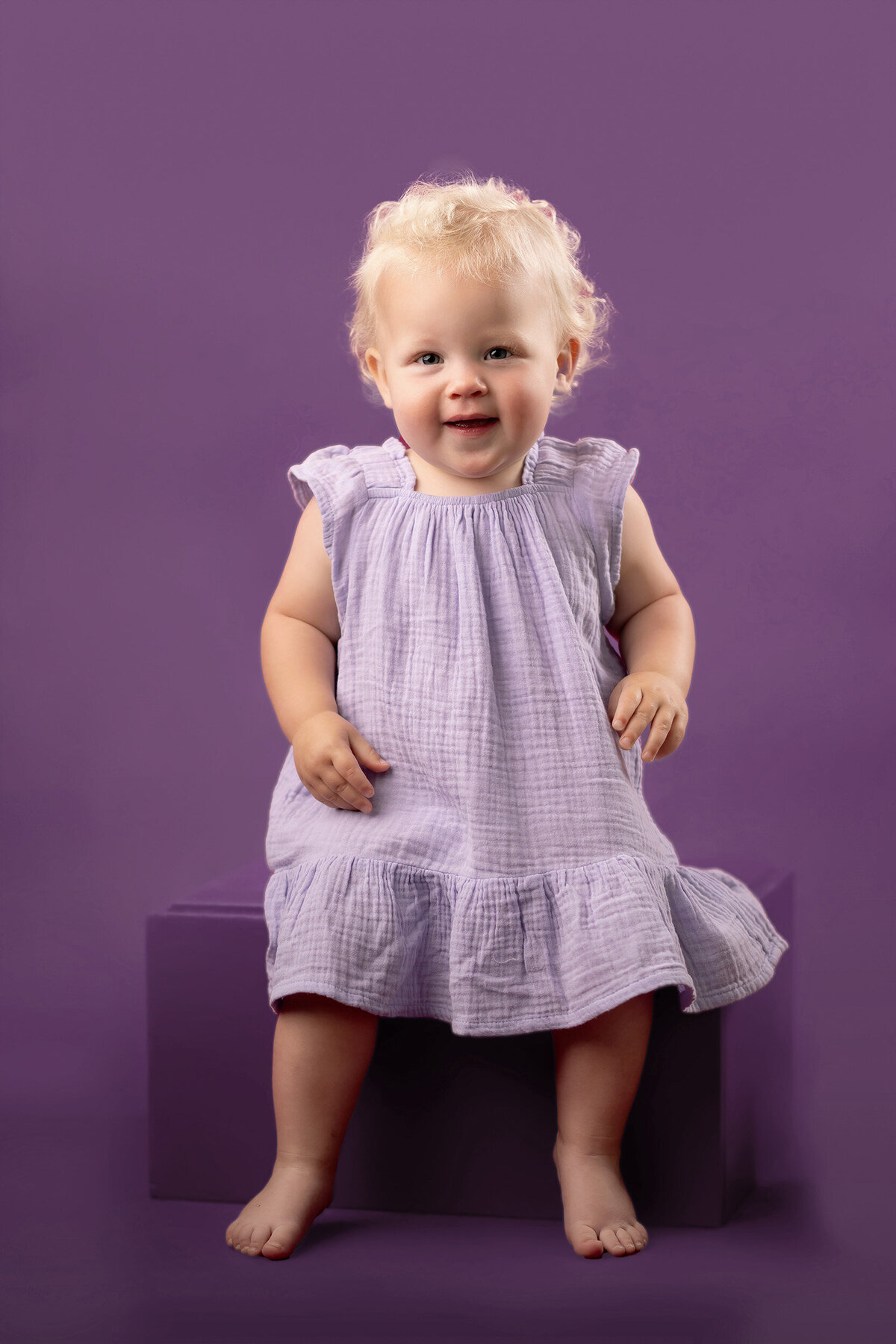 A toddler with curly hair sits barefoot on a block, wearing a lavender dress, against a matching purple background.