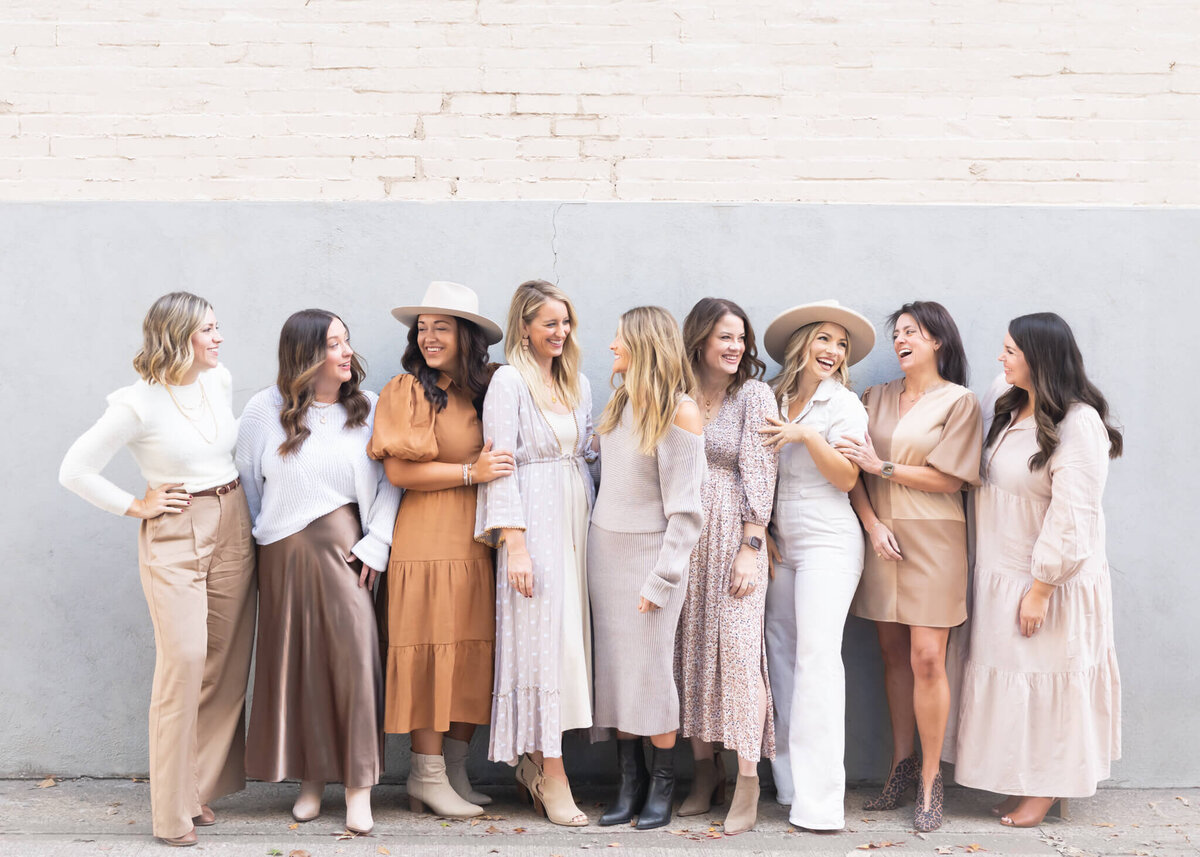 team photo of interior design team in downtown McKinney against a grey and white  wall, all team members wearing beige, white, tan, brown