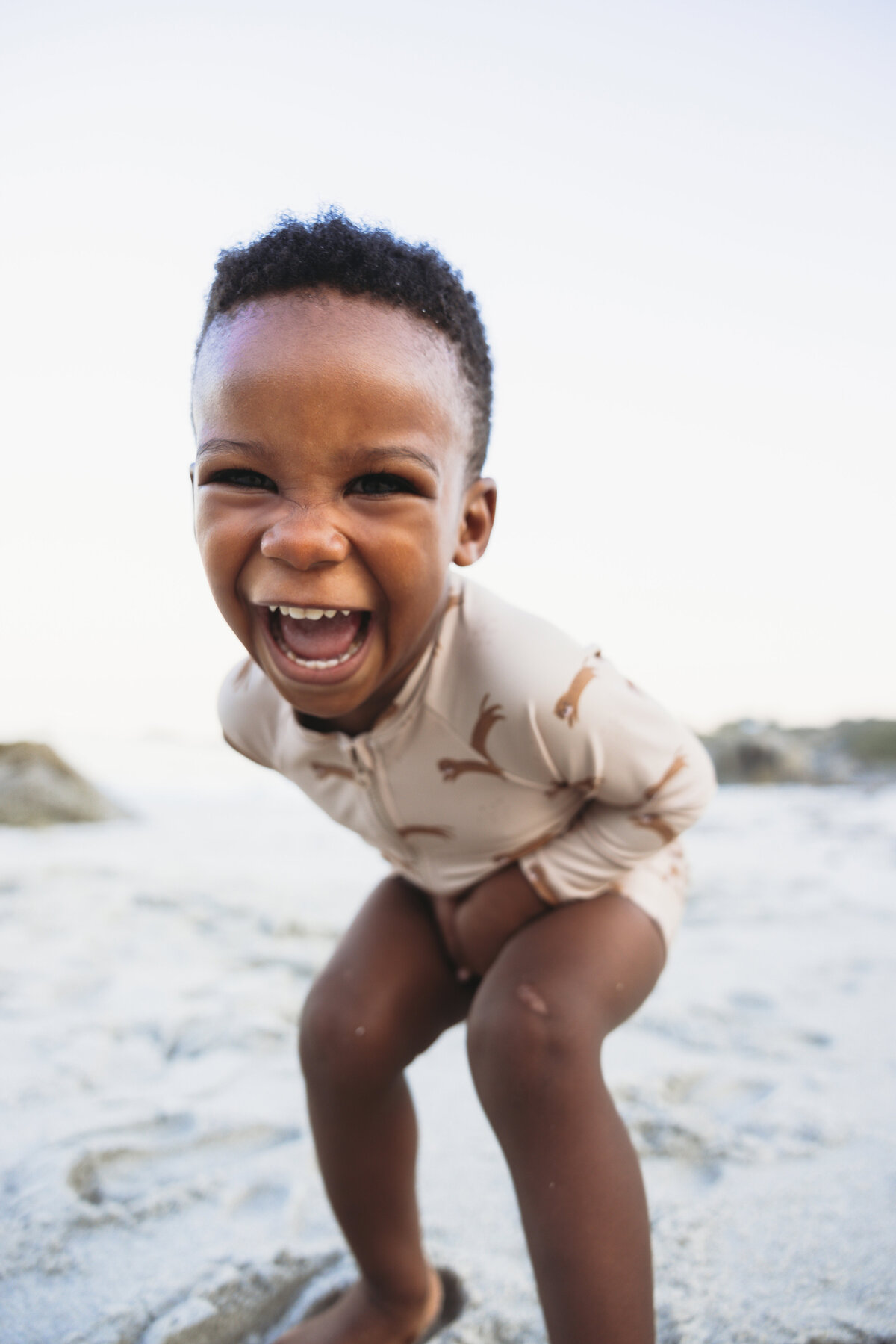 Laughing young boy in a beige UV swimsuit with cheetah print, bending forward playfully on a sandy beach in Cape Town.