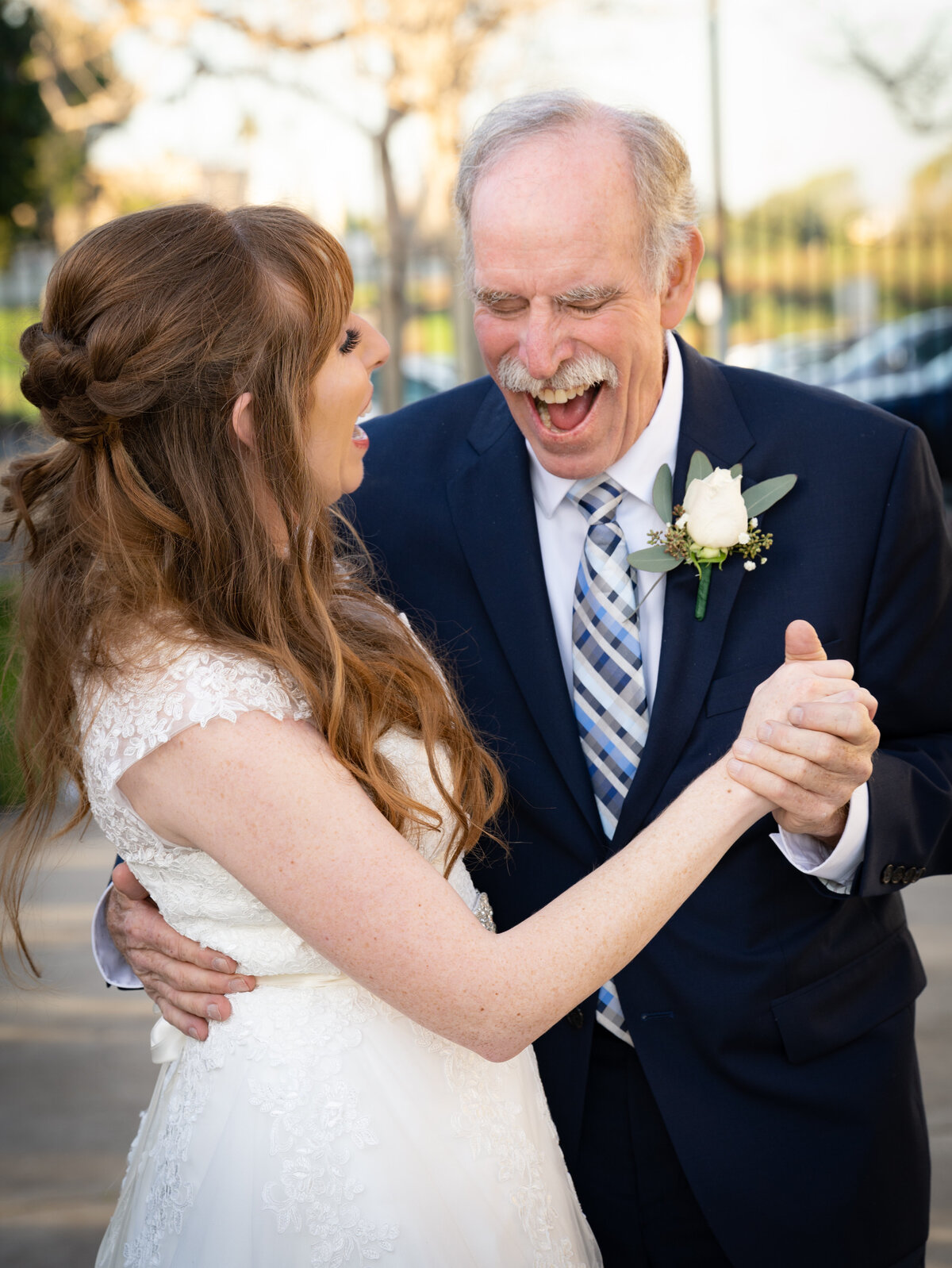 Elderly father in a navy suit with white-rose boutonniere holds red-headed daughter and laughs heartily with her during their special dance at her wedding. Photo by SAVI Photography - Photographer in Riverside