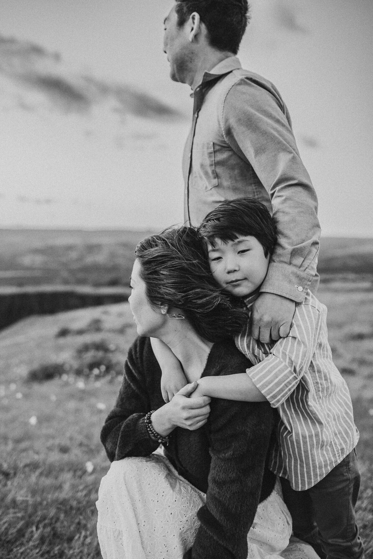 Black and white photo of a young boy holding his mom's hand while he rests on her back. The dad has his hand on his son's shoulder. 
