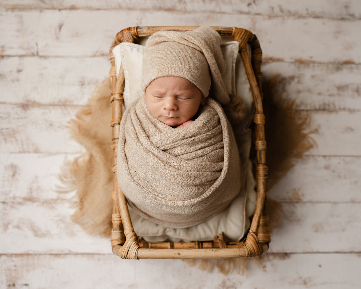 Newborn baby boy swaddled in warm beige tones with a matching sleepy cap, posed in a woven basket on a rustic white wood backdrop. Captured by Inland Empire newborn photographer Kaitlyn Dawn Photography.