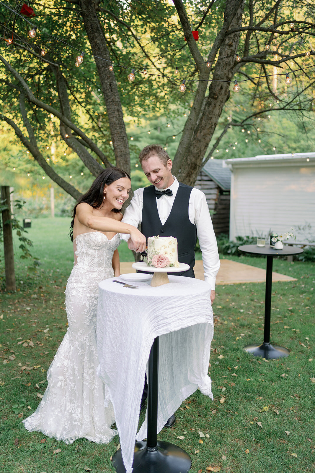 Bride and groom cutting their wedding cake outdoors under string lights at Glasshouse Community during their summer wedding.