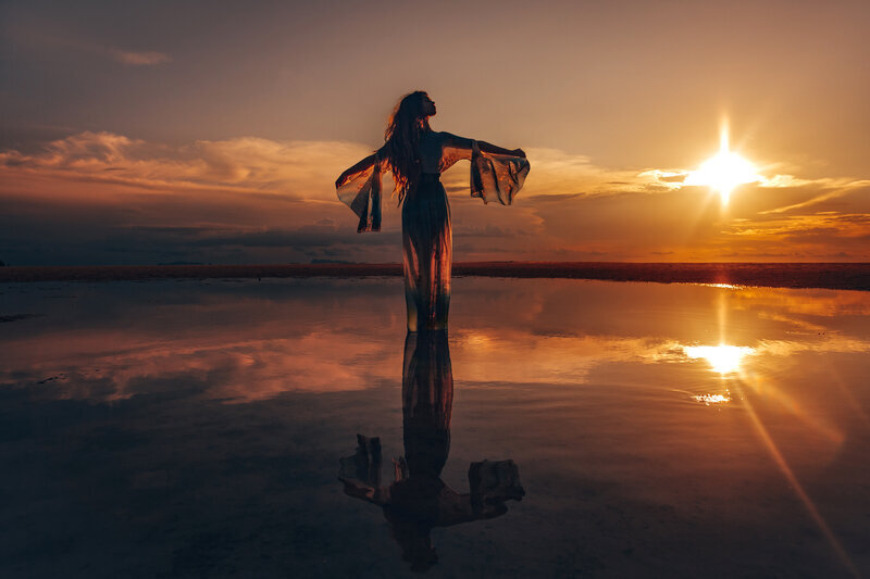Photo of an Eclectic witch in a long black dress with long fringed sleeves dancing at the water's edge beneath a beautiful blue sky with clouds
