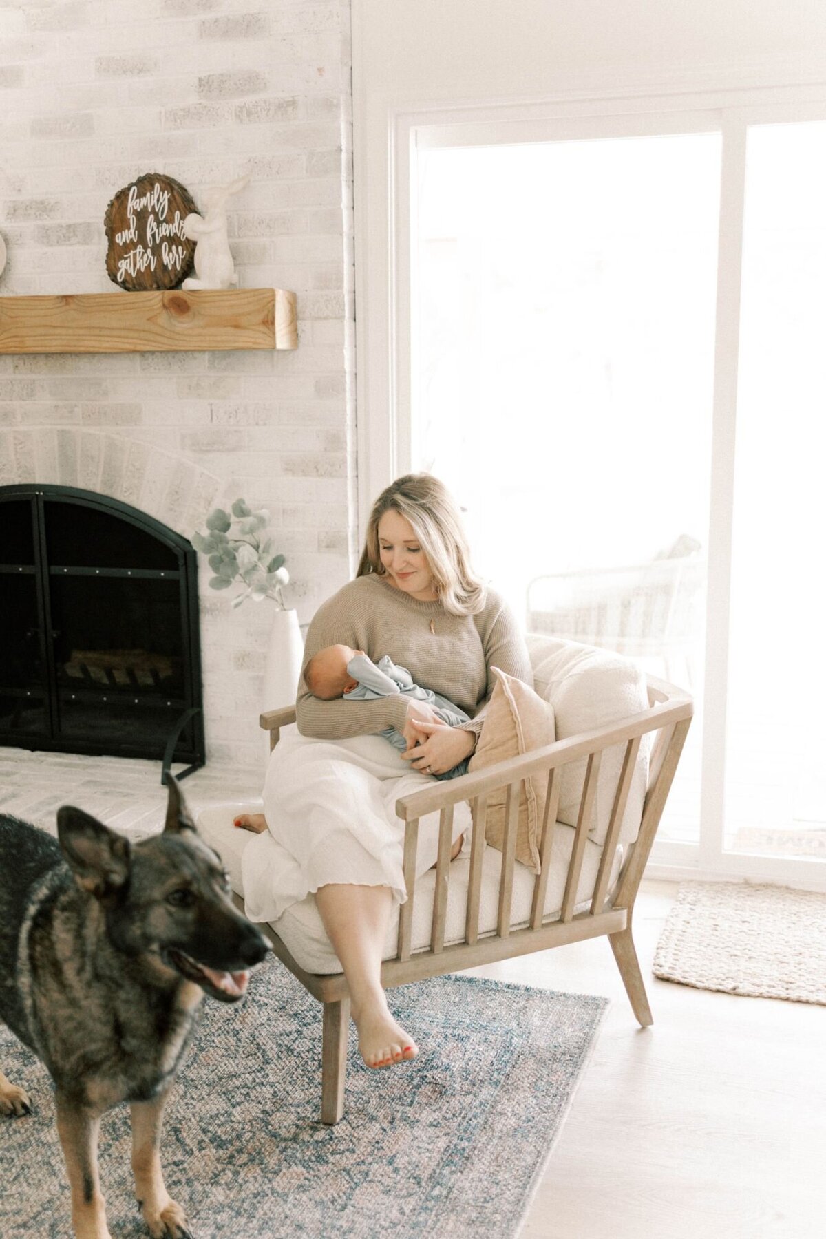 Woman sits on chair with baby and dog standing nearby.