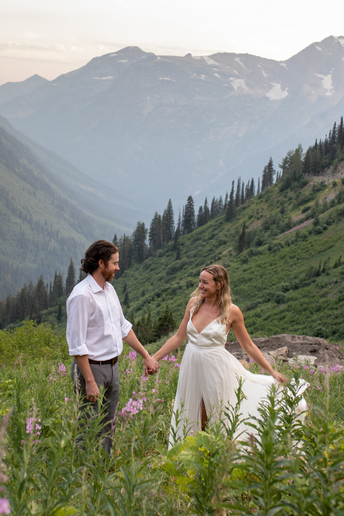 A bride and groom stand holding hands in a field of flowers in Glacier National Park.