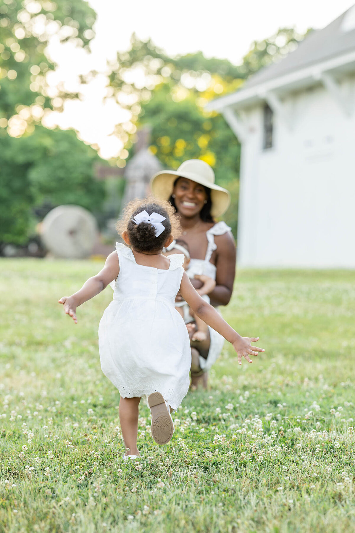 Toddler with both of her arms back run towards woman who is kneeling down in a clover field of grass