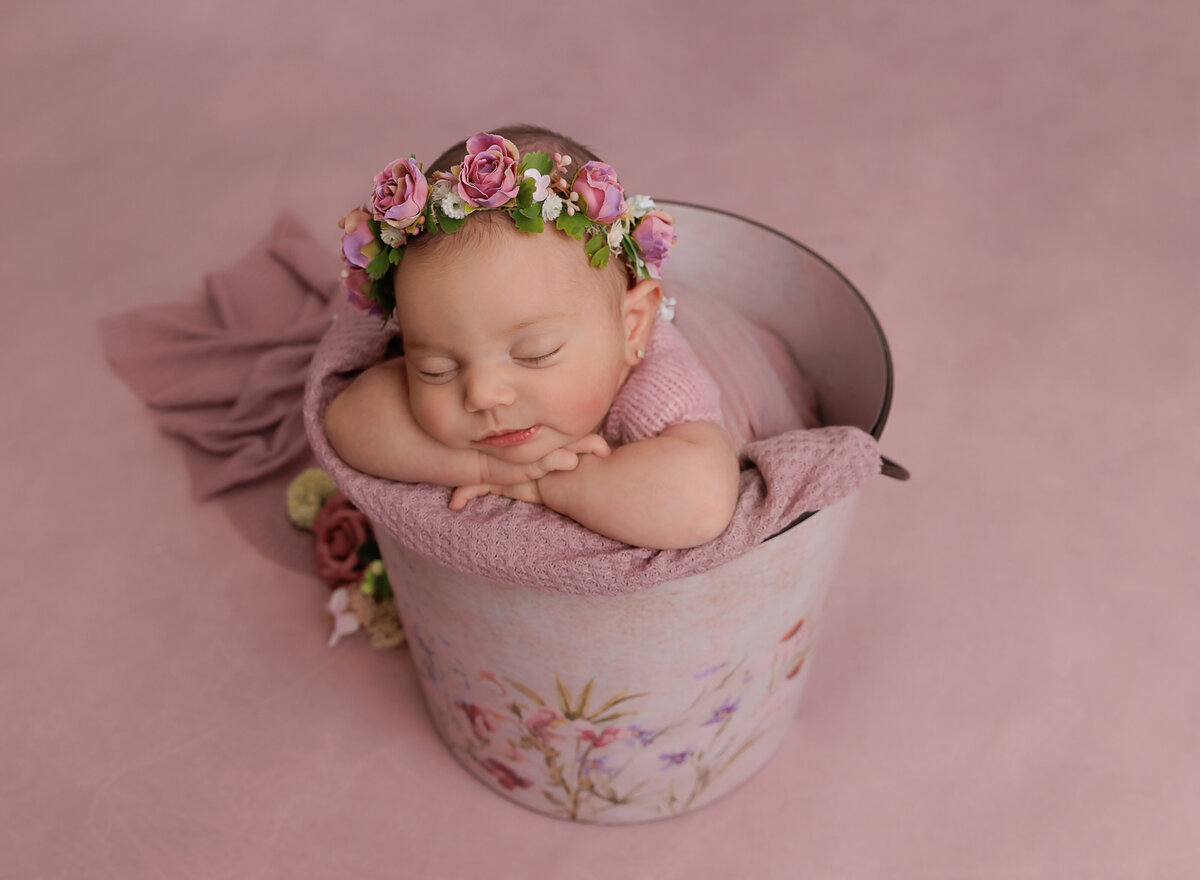 Baby girl is sleeping in a bucket for her newborn photoshoot. Baby's hands are folded underneath her chin and she is wearing a pink floral crown. Captured by top Brooklyn newborn photographer Chaya Bornstein