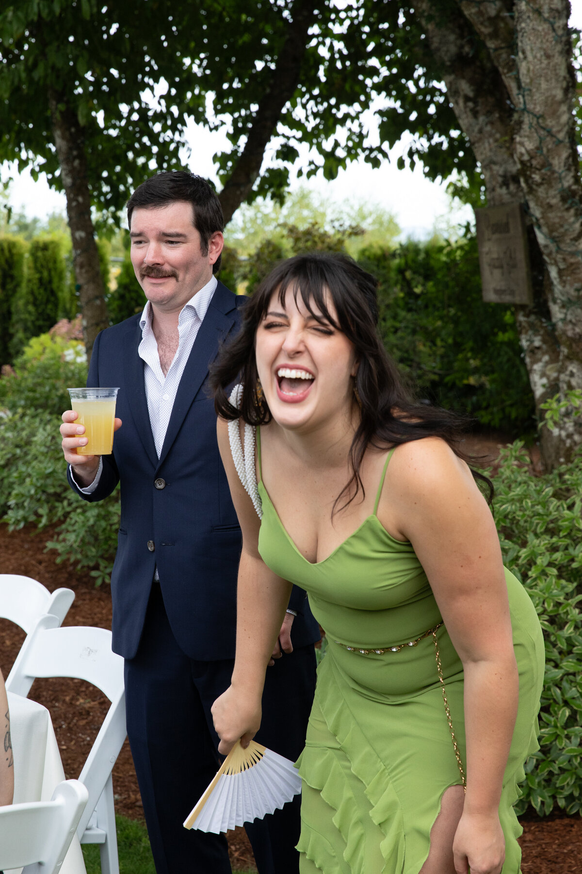 Guest in green dress laugh at a wedding reception while husband  stands nearby with drink
