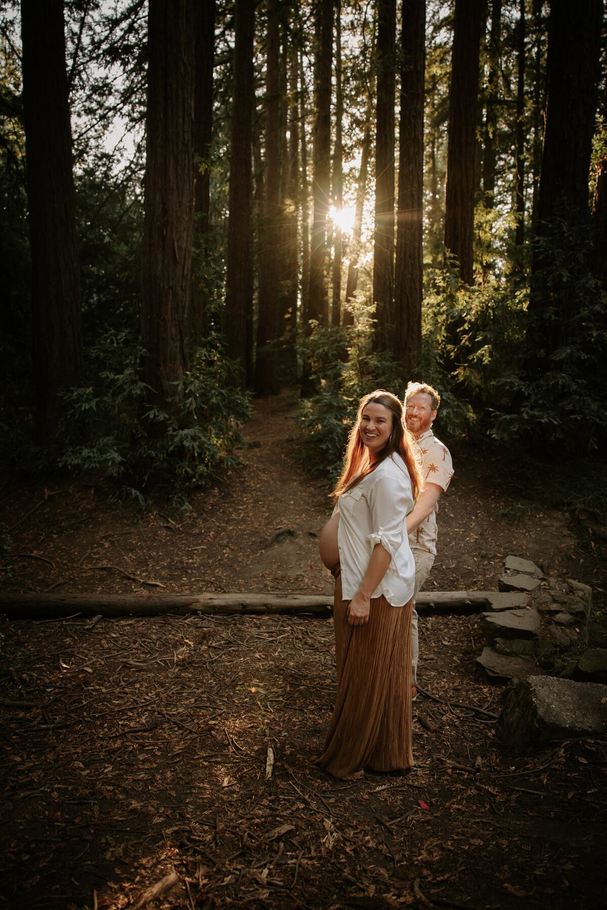 Pregnant woman and partner look on during sunset in the redwood grove