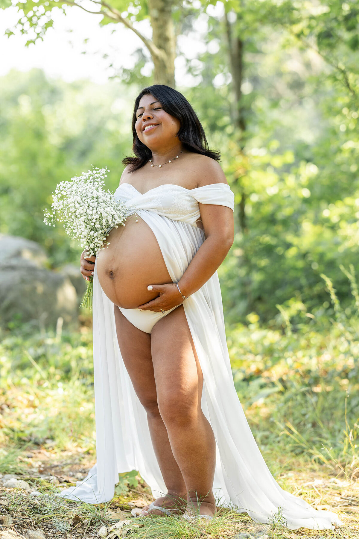 Expecting mom stands smiling with her eyes closed, hand on bump as she holds baby breath flowers with one hand during her maternity session.