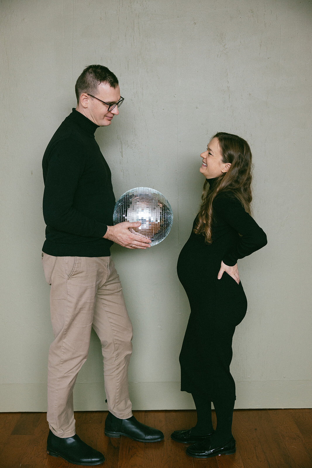 Man and woman laugh while looking at each other as man holds disco ball representing womans pregnant belly. 