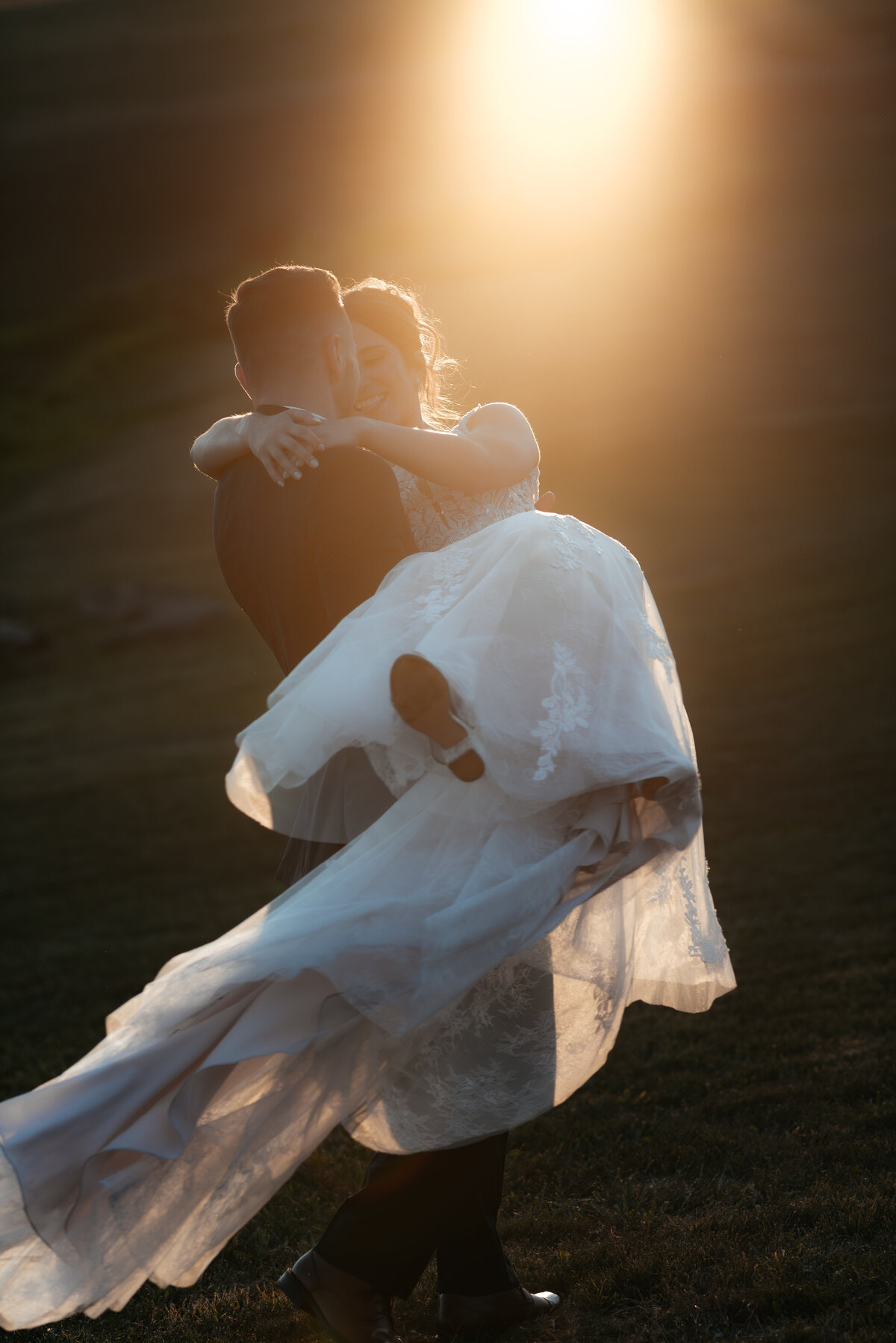 The groom carries the bride with her long train swinging behind her as they laugh candidly during a sunset session.