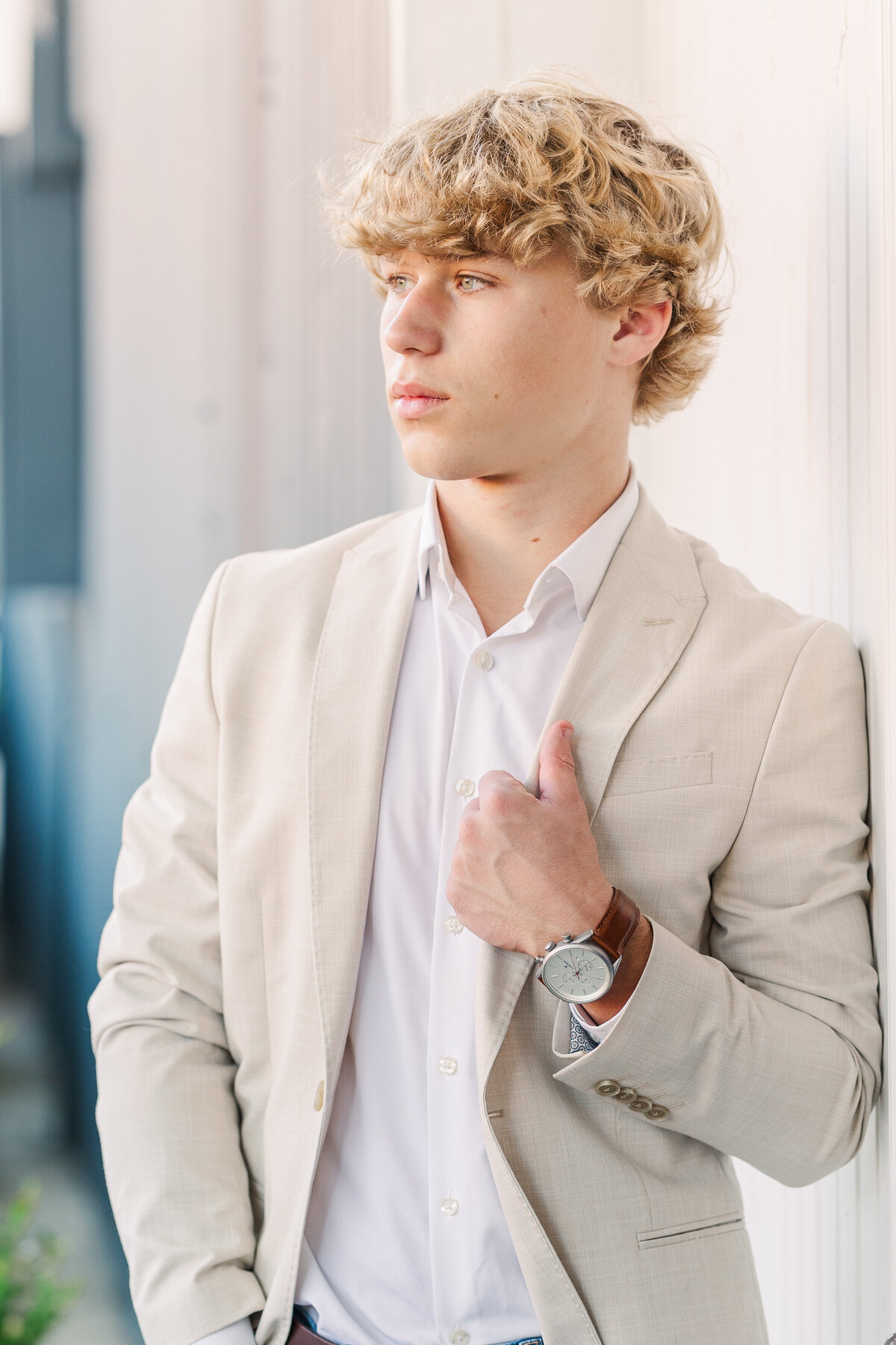 Editorial-style senior portrait of a young man in a light tan suit jacket and white dress shirt, looking off-camera with a confident expression. He wears a silver watch with a brown leather band and leans against a light-colored industrial wall in natural light.