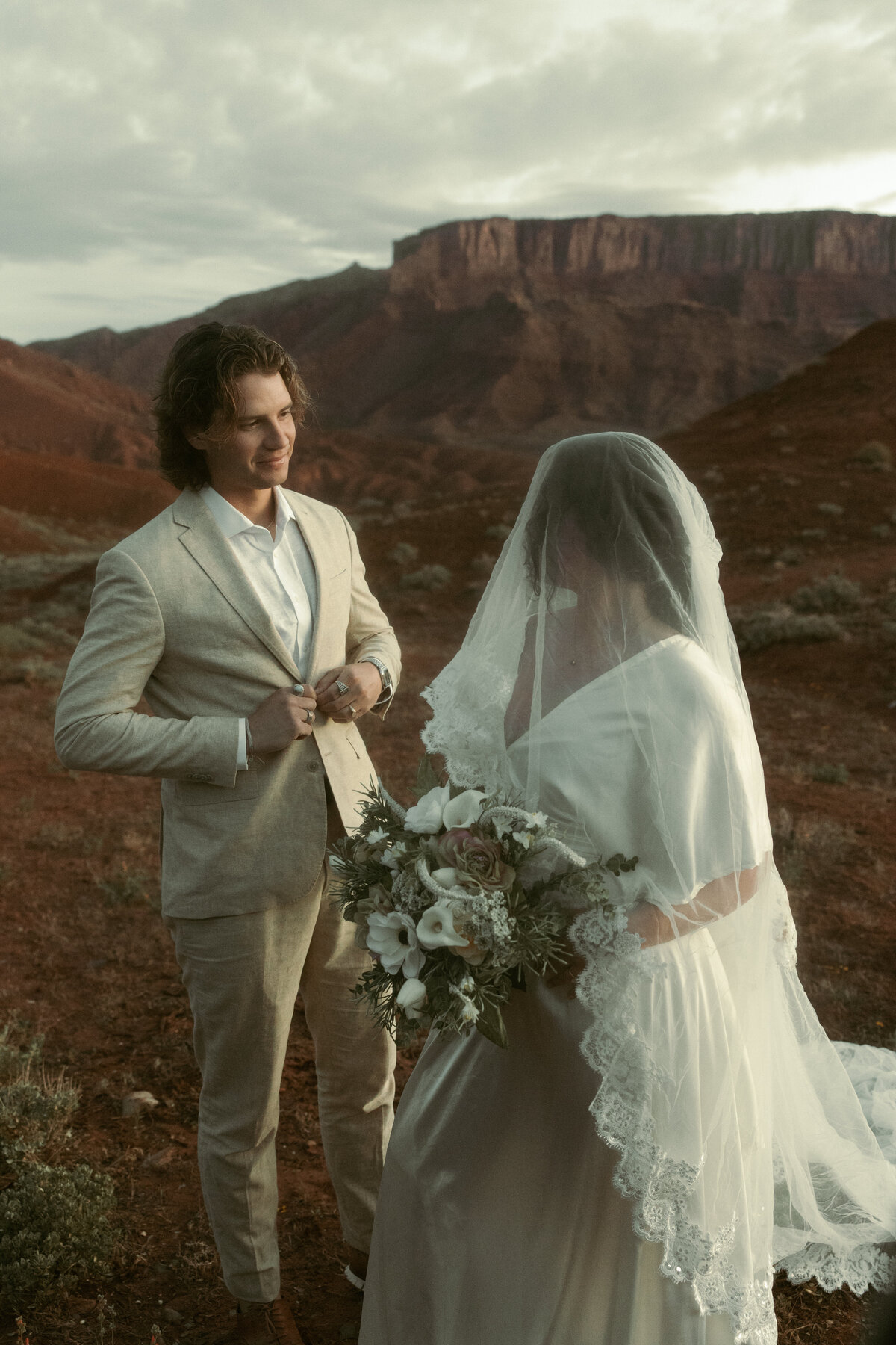 A bride in a lace-trimmed veil and a bouquet stands near a groom adjusting his suit, set against dramatic red rock formations.
