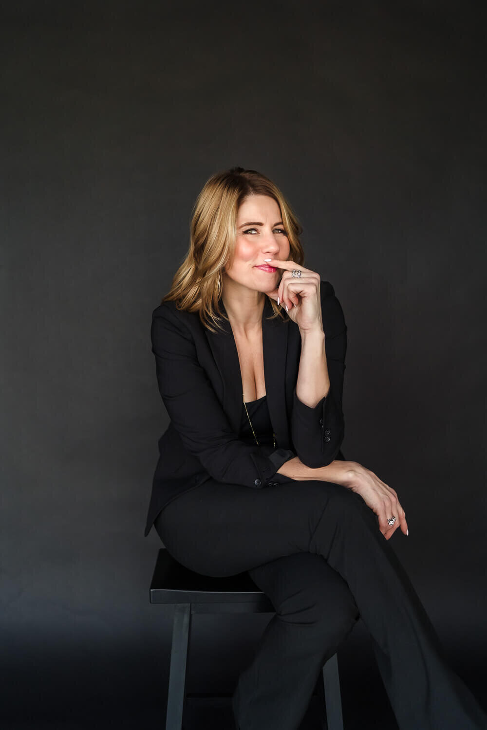 Woman in black suit sitting on stool with hand at chin during headshot session