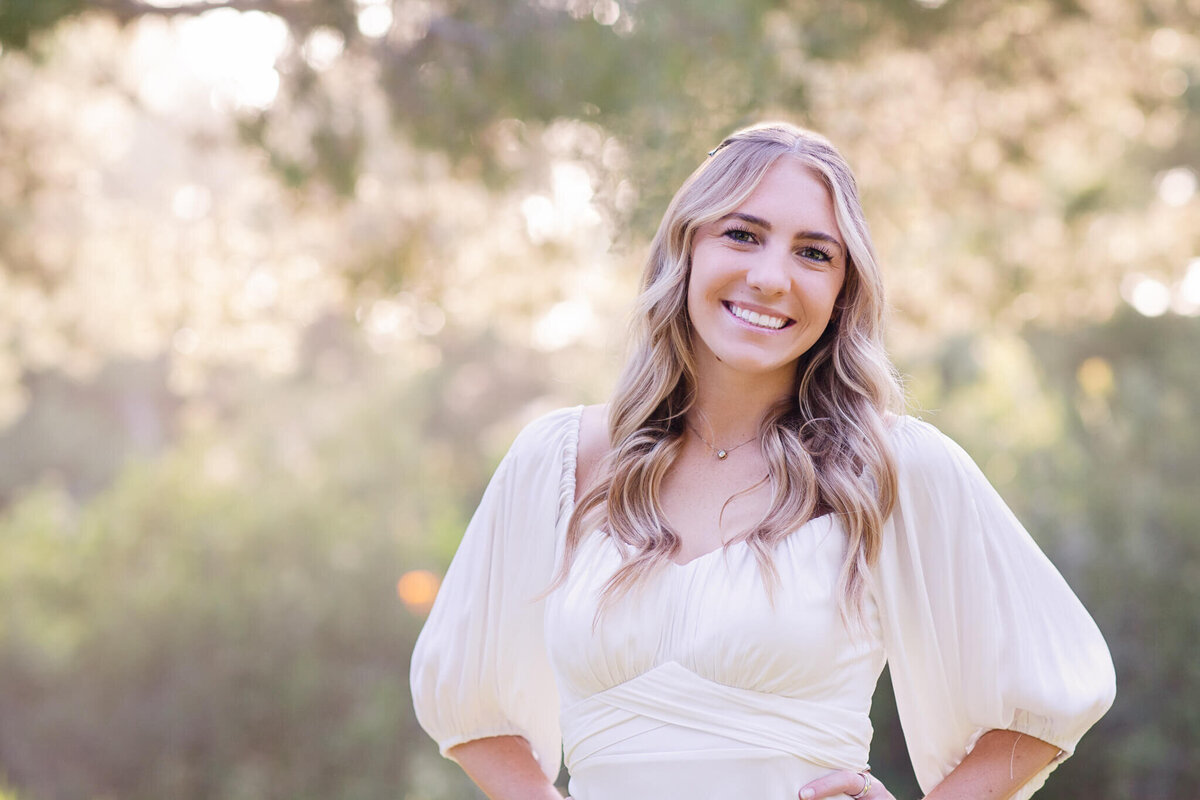 A woman with long blonde hair smiles at the camera, standing outdoors. She is wearing a white dress with puffed sleeves. The background is blurred with green foliage and soft, dappled sunlight.