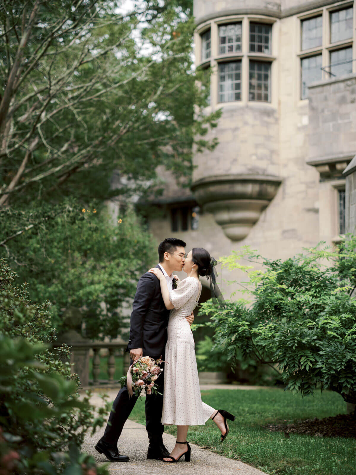 The engaged couple is kissing outside Coe Hall at Planting Fields Arboretum, NY, amidst lush greenery. Image by Jenny Fu Studio