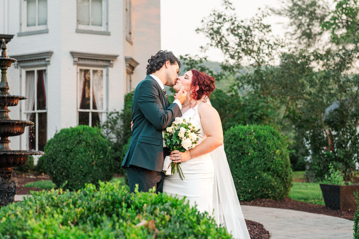 Bride and groom kissing behind a bush in front of Richwood on the River mansion