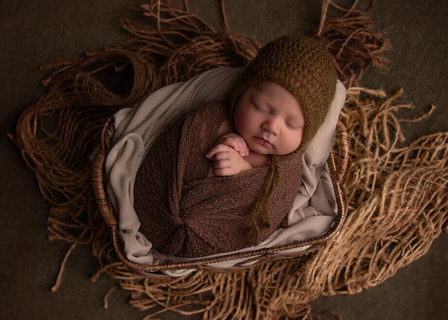 This baby boy has been wrapped in a brown cheesecloth wrap with a twist in front.  He has been positioned in a brown basket on top of a textured decorative throw and a forest green backdrop