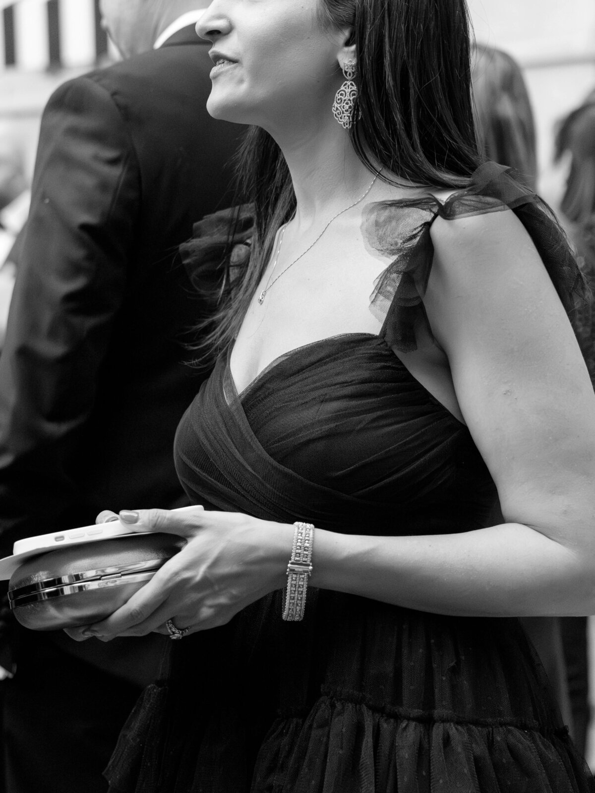 A woman in an elegant black dress holds a clutch purse at a formal event. She wears statement earrings, a necklace, and a bracelet. The background shows multiple people in formal attire. The image is in black and white.