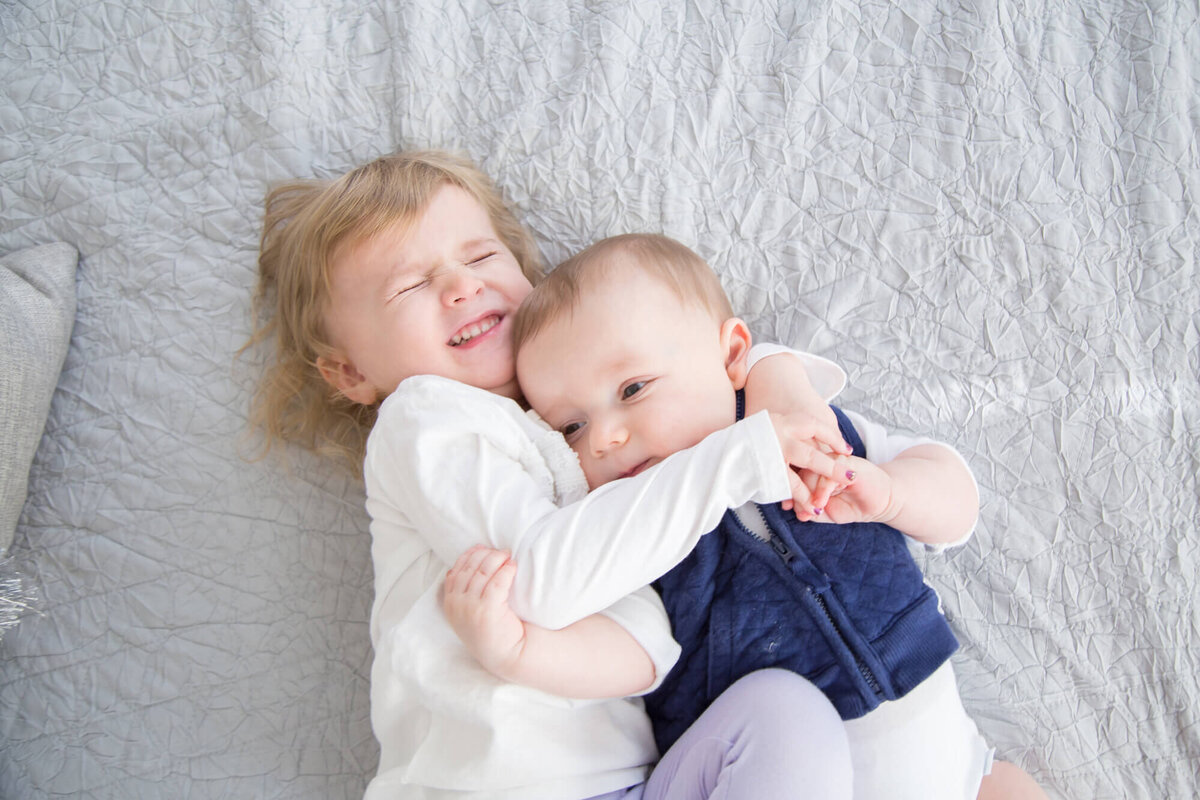 Toddler girl hugging infant brother tightly on a bed