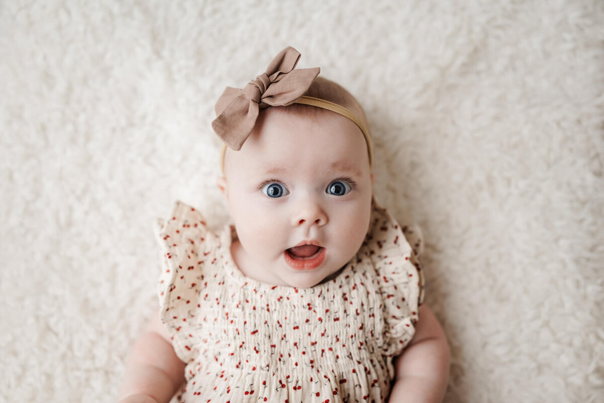A baby, captured by a portrait photographer from Bloomington, Illinois, wears a beige bow headband and a cream dress with tiny red patterns. Lying on a soft, white textured surface, the baby has a surprised expression with wide eyes and slightly open mouth.