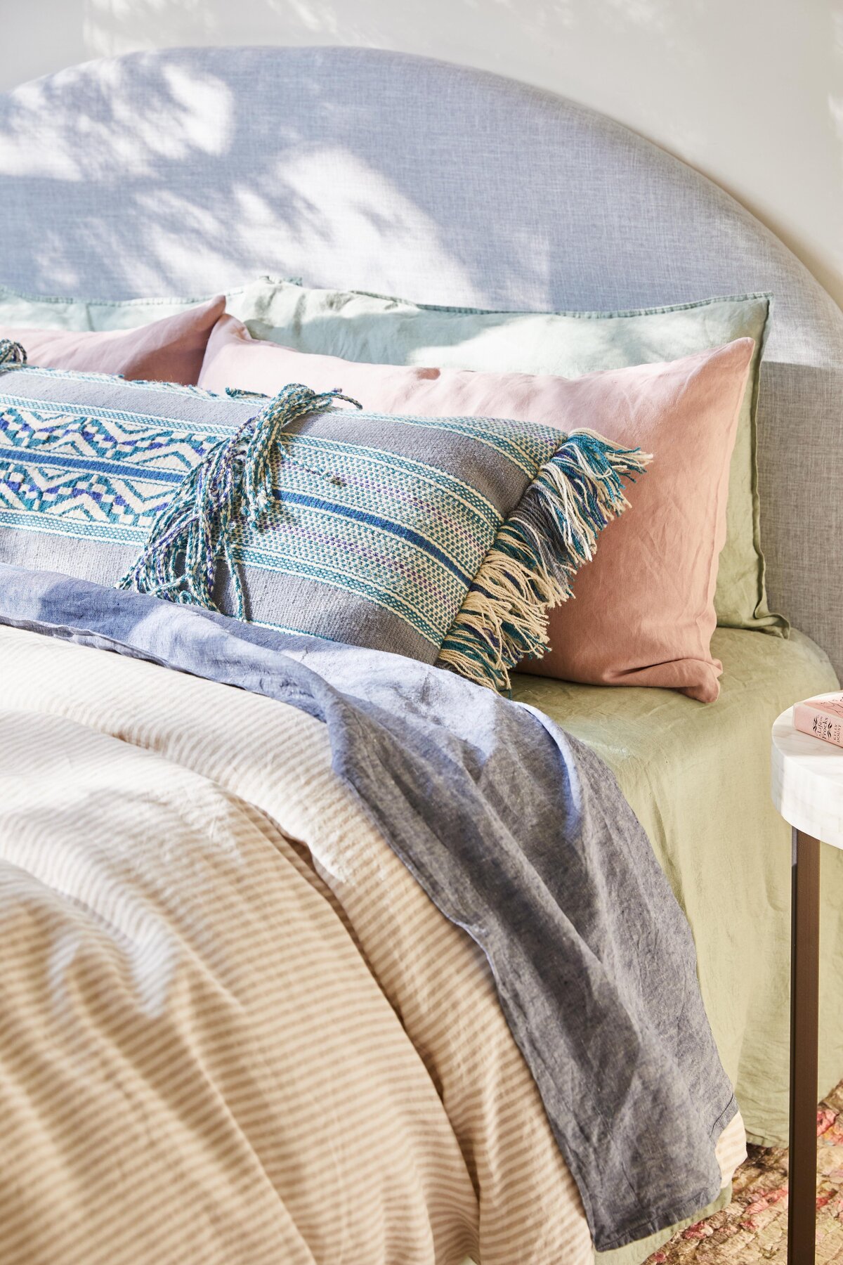 Elwood bedroom detail with a light‑grey curved headboard, sage and blush linen pillows and a blue‑striped tasselled lumbar cushion for texture and colour.
