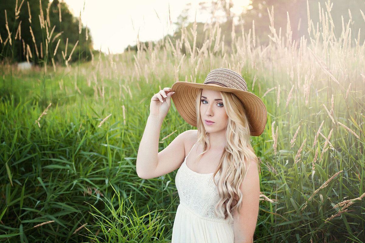Long blonde haired young woman wearing floppy brimmed hat standing in open field with tall grass captured in the summer time with Studio 64 Photography Akeley, Minnesota.