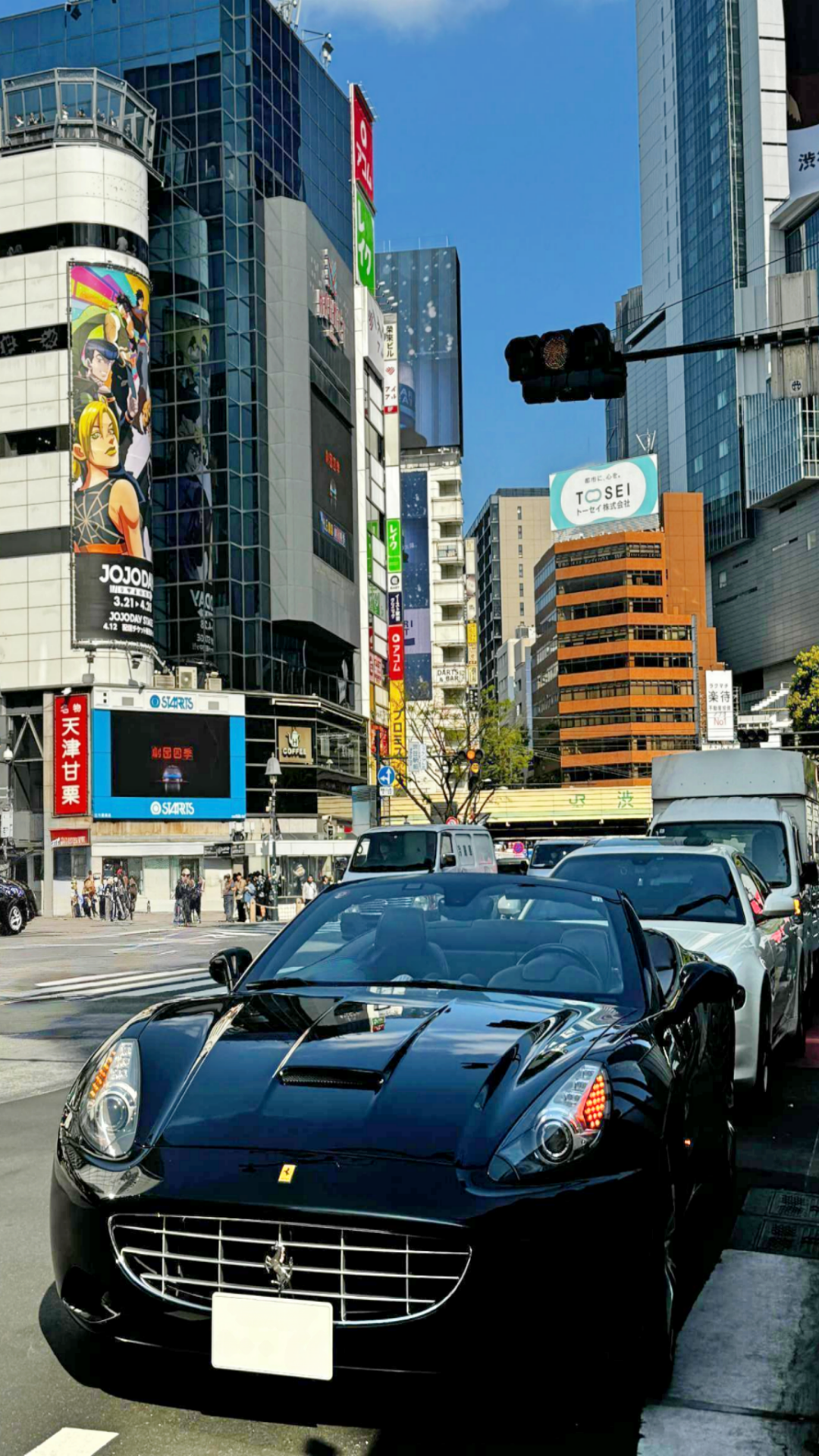 A sleek black Ferrari convertible driving through the bustling streets of Shibuya, Tokyo, surrounded by modern buildings, LED billboards, and city traffic on a sunny day.