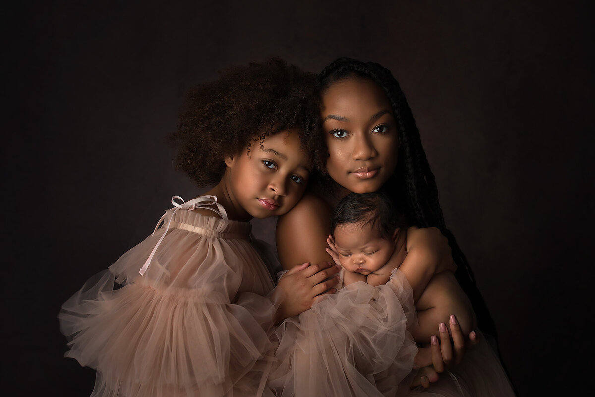 Two sisters in blush tulle dresses holding their newborn sibling against a dark backdrop, captured by Dallas newborn photographer.