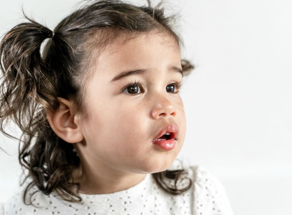 A close-up color portrait of a little girl with an expressive face. She has curly brown hair styled in two pigtails and is looking off to the side with her mouth slightly open. She is wearing a white dress with eyelet cutouts, and the background is a simple white.