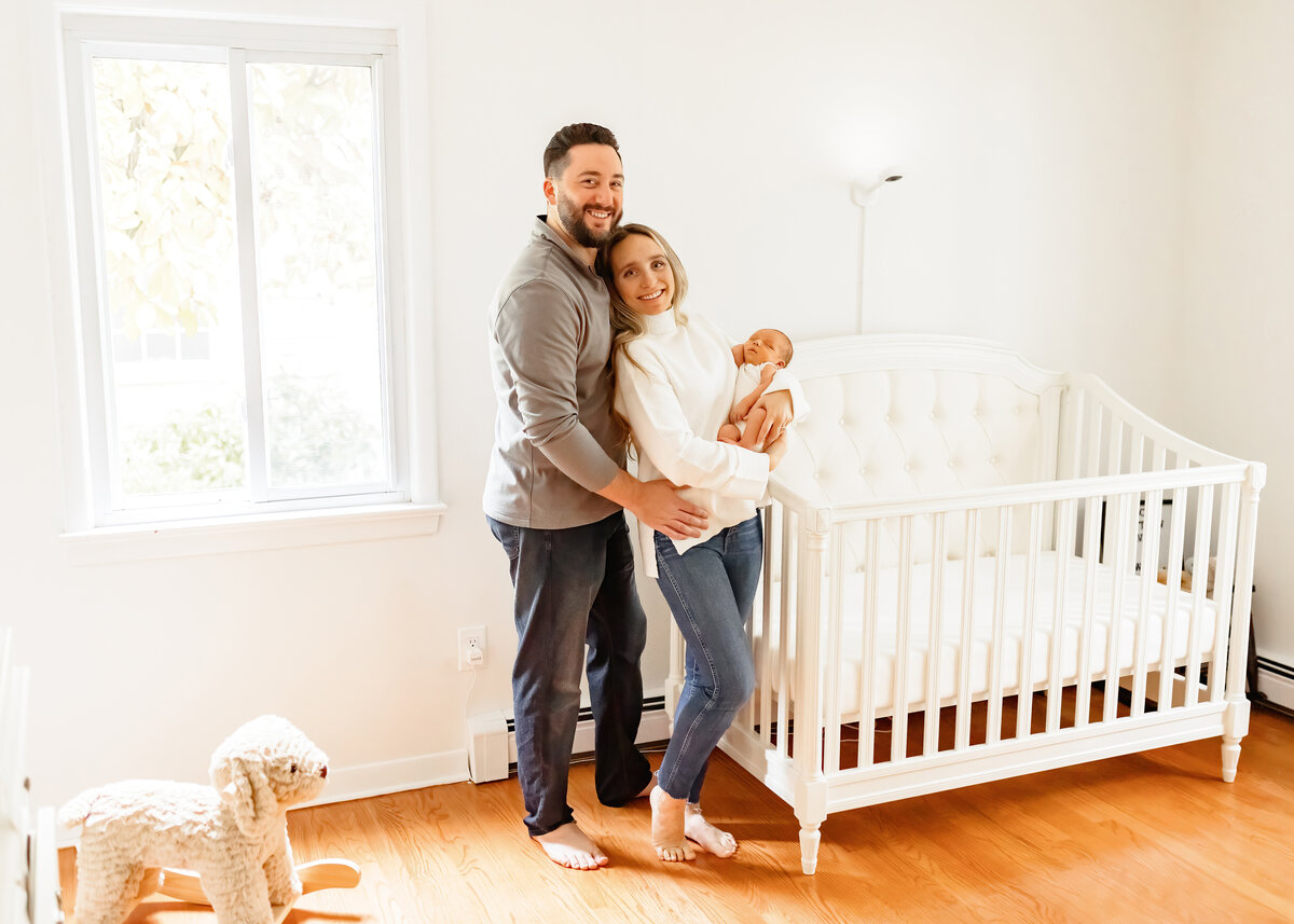 parents standing next to a crib holding their newborn baby and smiling 