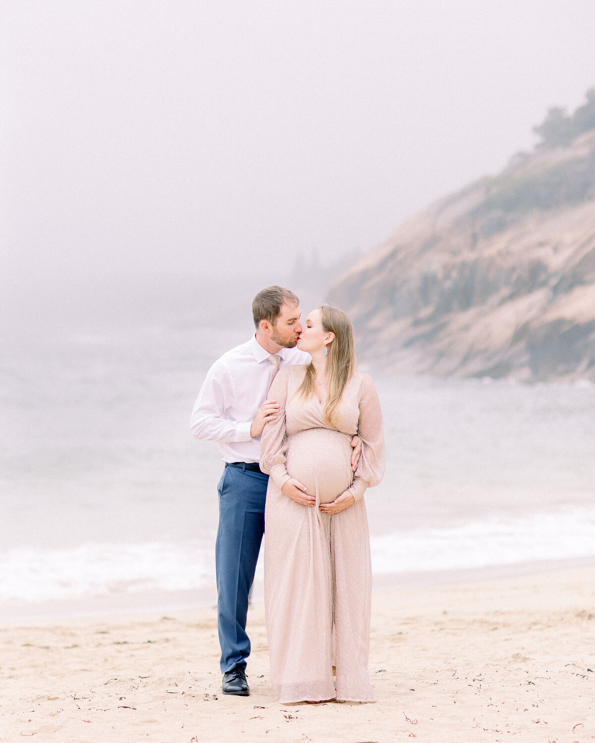 Pregnant mom and dad kissing on a misty beach with mountains emerging from the fog in the background , by Wellesley, MA maternity photographer Fieldstone Studio.