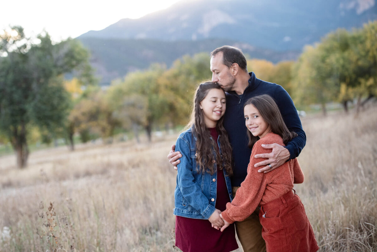 A father kisses his daughter's head while hugging the other in a park field at sunset for a Denver family photographer