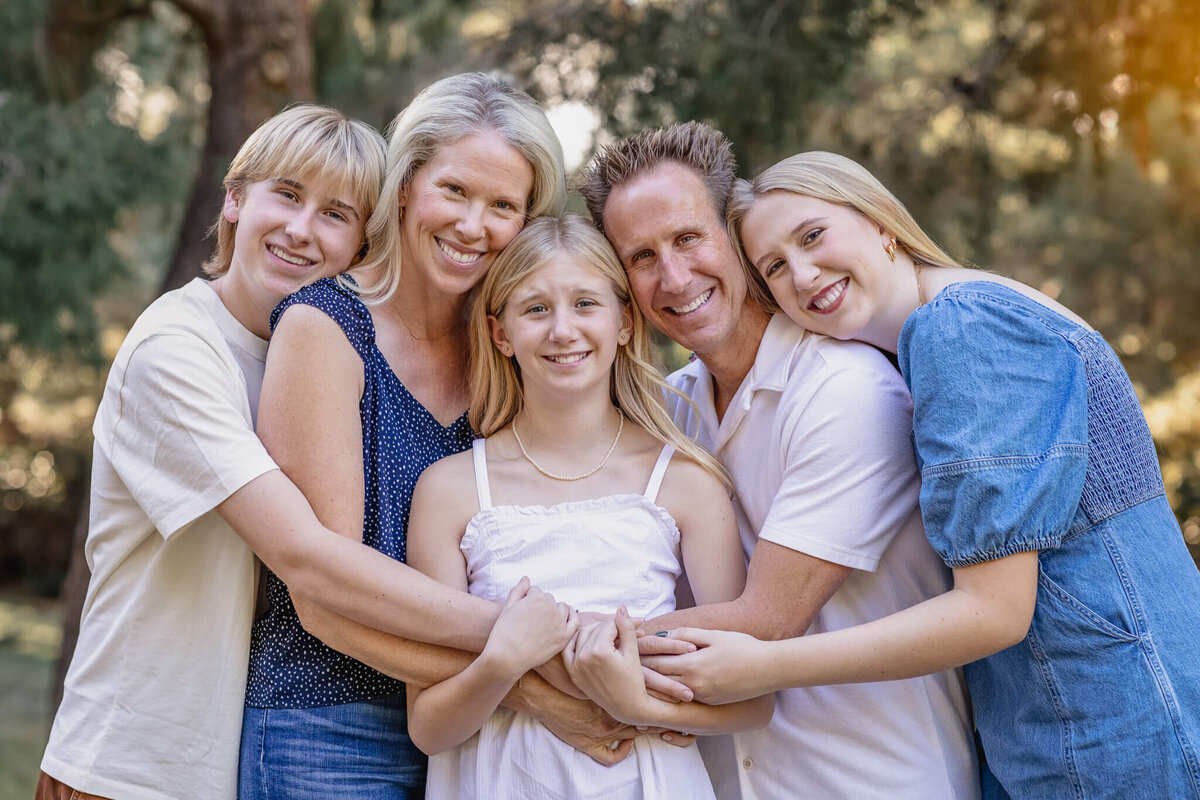 A family of five with blonde hair hugging each other with their arms around each other and their heads together in a park.